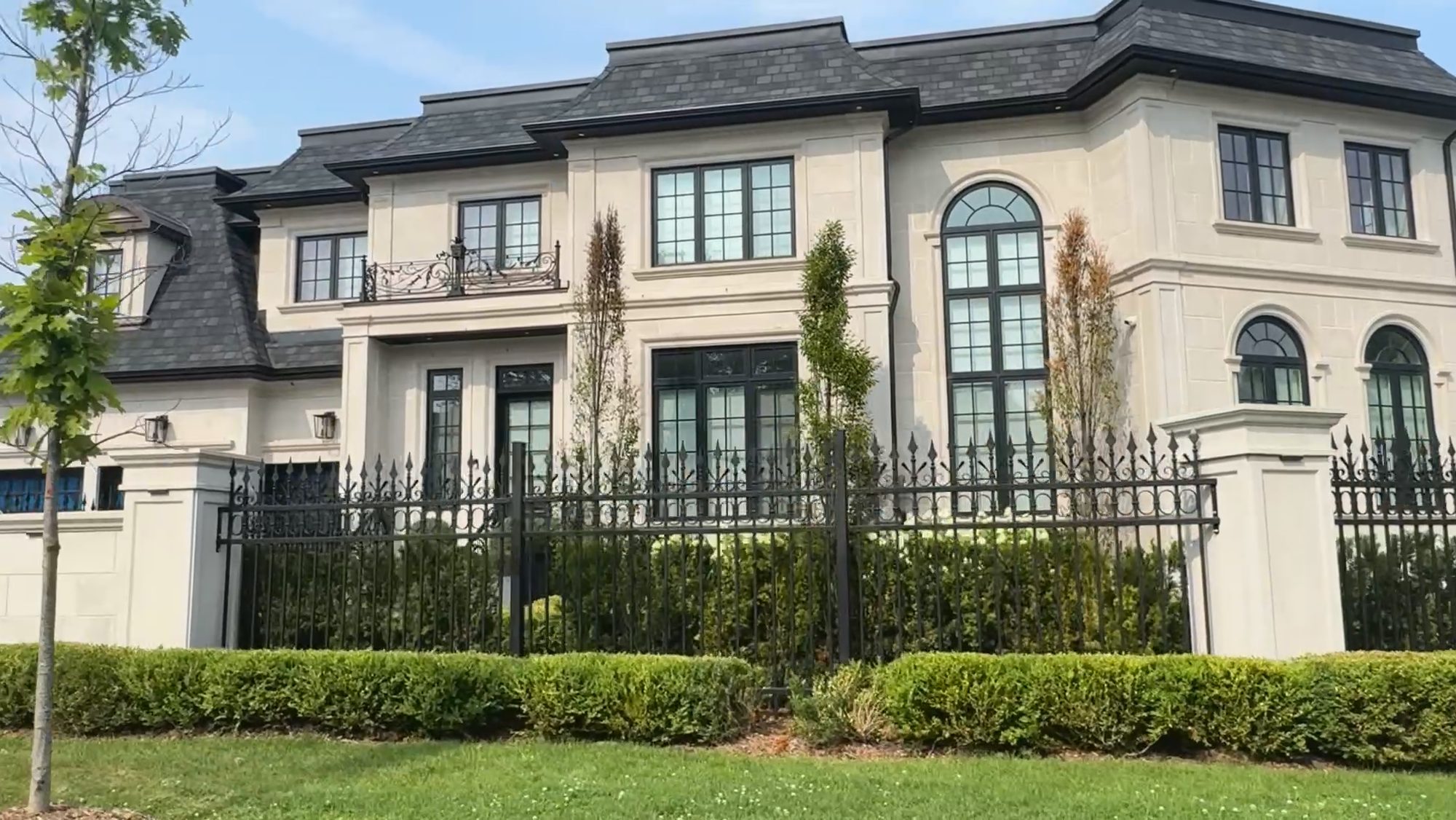 A large stone-and-stucco house behind a clipped hedge and iron fence