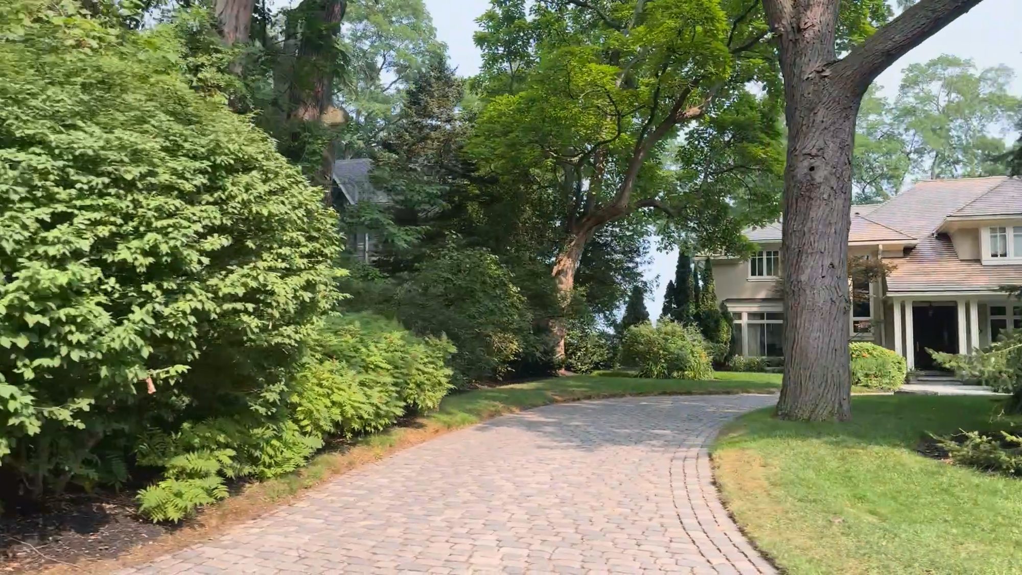 A brick-paved driveway curving between mature trees toward a large house